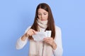 Young unwell dark haired woman wearing white sweater and scarf, holding recipe in hand, reads it with astonished expression,