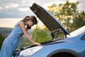 Young upset woman driver talking on mobile phone near a broken car with open hood inspecting engine having trouble with her