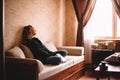 Young upset woman holding book looking through window while sitting on sofa in living room Royalty Free Stock Photo