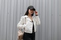 Young urban brunette woman in a fashionable white leather jacket in vintage jeans in a t-shirt in a stylish black cap Royalty Free Stock Photo