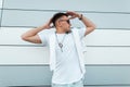 Young urban hipster man with stylish hairstyle in a fashionable white T-shirt in vintage sunglasses posing outdoors near a modern Royalty Free Stock Photo