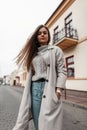 Young urban woman model in a vintage trench coat in blue jeans in a knitted stylish sweater is resting standing on the street near