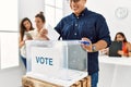 Young voter man smiling happy putting vote in voting box standing by ballot at electoral center Royalty Free Stock Photo