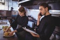 Young waiter talking on smartphone while waitress preparing food in commercial kitchen