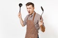 Young warlike man chef or waiter in striped brown apron, shirt holding black ladle or kitchen spoon, spatula isolated on