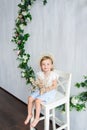 A young warm smiling girl is sitting on the wooden white chair near the wall with flowers wreath.