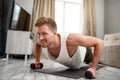 Young well-built man go in for sports in apartment. Strong muscular guy stand in plank position using dumbbells and look