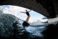 Young wet woman in black swimming vest rides down the river wave on wake board