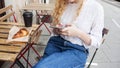 Young white blond woman having a break and holds smartphone in hand. Street veranda table in cafe with cardboard paper cup of Royalty Free Stock Photo