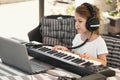 A young white Caucasian girl wearing headphones and playing a keyboard connected to a laptop personal computer