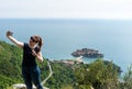 Young white girl making a selfie in the mountain and the adriatic sea in the background and Sveti Stefan.Woman with smart phone in