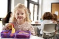 Young white schoolgirl sitting at a table smiling and holding an apple in a kindergarten classroom during her lunch break, close u