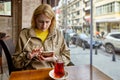 Young white woman sits at table by window in cafe with Turkish glass of tea in front of her and reads messages on mobile phone