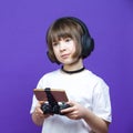 Young Winsome Girl in White Shirt Posing With Gamepad Joystick And Wireless Headphones While Using Smartphone For Gaming With Calm