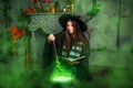 A young witch in a black hat and mantle makes a potion in a black cauldron with a magic wand and a book. A girl sits on the floor Royalty Free Stock Photo