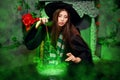 A young witch in a black hat and mantle makes a potion in a black cauldron with a magic wand and a book. A girl sits on the floor Royalty Free Stock Photo