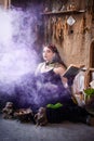 A young witch with a book in her hands cooks a potion in a large black cauldron, emitting magical purple smoke