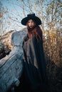 A young witch with pale skin and black lips, wearing a black hat, dress and cloak. In autumn, against the background of a fallen t