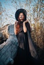 A young witch with pale skin and black lips, wearing a black hat, dress and cloak. In autumn, against the background of a fallen t