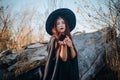 A young witch with pale skin and black lips, wearing a black hat, dress and cloak. In autumn, against the background of a fallen t