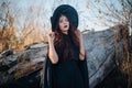 A young witch with pale skin and black lips, wearing a black hat, dress and cloak. In autumn, against the background of a fallen t