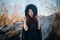 A young witch with pale skin and black lips, wearing a black hat, dress and cloak. In autumn, against the background of a fallen t