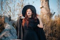 A young witch with pale skin and black lips, wearing a black hat, dress and cloak. In autumn, against the background of a fallen t