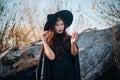 A young witch with pale skin and black lips, wearing a black hat, dress and cloak. In autumn, against the background of a fallen t