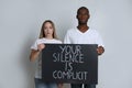 Young woman and African American man holding sign with phrase Your Silence Is Complicit on grey background. Racism concept