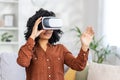 Young woman alone sitting on couch in living room at home, African American woman using virtual reality glasses, for Royalty Free Stock Photo