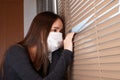 A young woman in an antibacterial mask looks out the window from the house to the street, sits in the apartment
