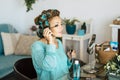 Young woman applying mascara in eyelashes while looking at mirror in living room at home. Caucasian woman using curlers for hair