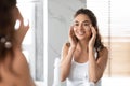 Young Woman Applying Moisturizing Facial Cream Looking In Mirror Indoor