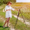 Young woman artist at the easel painting landscape outdoors. Girl-artist working on the plein air Royalty Free Stock Photo