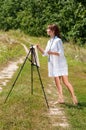 Young woman artist at the easel painting landscape outdoors. Girl-artist working on the plein air Royalty Free Stock Photo