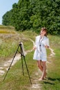 Young woman artist at the easel painting landscape outdoors. Girl-artist working on the plein air Royalty Free Stock Photo