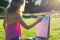 A young woman artist holds a brush and paints a picture on an easel iof the sunset