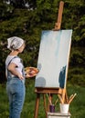 A young woman artist holds a brush and paints a picture on an easel in the rays of the sunset. The painter paints oil paintings in Royalty Free Stock Photo