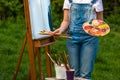 A young woman artist holds a brush and paints a picture on an easel in the rays of the sunset. The painter paints oil paintings in Royalty Free Stock Photo