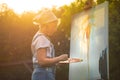 A young woman artist holds a brush and paints a picture on an easel in the rays of the sunset. The painter paints oil paintings in Royalty Free Stock Photo