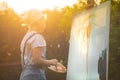 A young woman artist holds a brush and paints a picture on an easel in the rays of the sunset. The painter paints oil paintings in Royalty Free Stock Photo