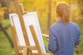 Young woman artist standing near an easel and looking at the sunset on the lawn, the girl is looking for inspiration for painting