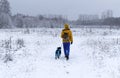 Young woman from the back in yellow winter clothes walking with a fluffy gray dog in a blue overalls in a winter snowy forest Royalty Free Stock Photo