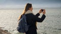Young woman with backpack on the rocky coast, taking photos of the cold winter sea on her smartphone. Perfect for tourism and Royalty Free Stock Photo
