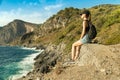 Young woman with backpack sit and rest on rock looking at the sea from rocky coast at sunset Royalty Free Stock Photo