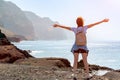 Young woman with backpack stand in front of ocean rocky shore . Girl near the sea spread her arms welcoming the sun on a Royalty Free Stock Photo