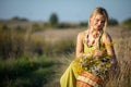 A young woman with a basket full of herbs crouches by a dirt road in the countryside and looks at them.