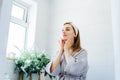 Young woman in bathrobe looking in mirror and doing manual self massage of face in the bathroom. Natural procedures for