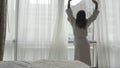 Young woman in bathrobe unveil curtains stretches arms and looks from the window