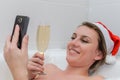 Young woman in the bathroom wearing a santa claus hat talks by video call on a smartphone and drinks champagne congratulating frie